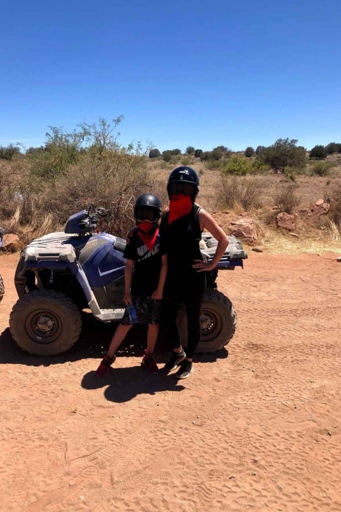mom and son standing in front of a blue ATV wearing black clothes, black helmets on top of head, black goggles over eyes and a red bandanna around face 