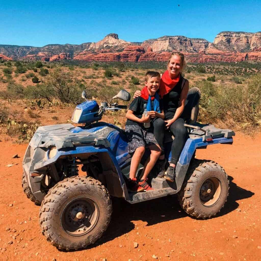 mom and son sitting on a blue atv in desert with red rock hills in the background