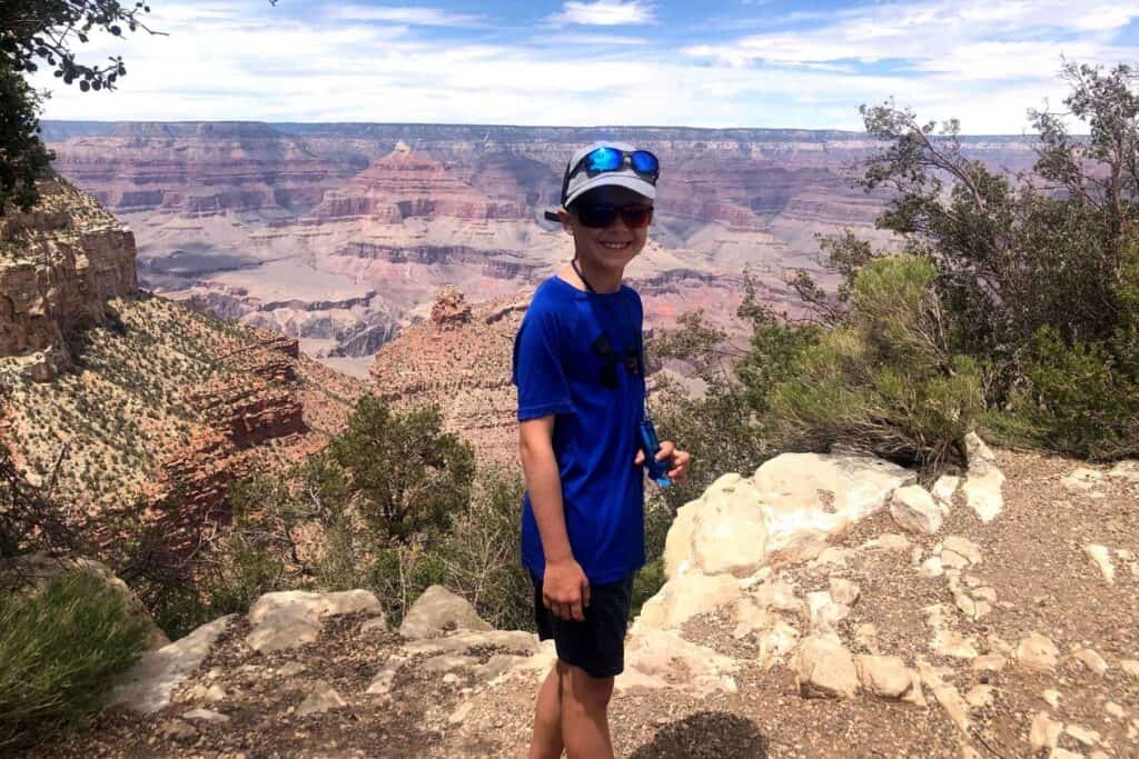boy standing in a blue shirt with a hat and glasses on with a red rock mountain valley behind him