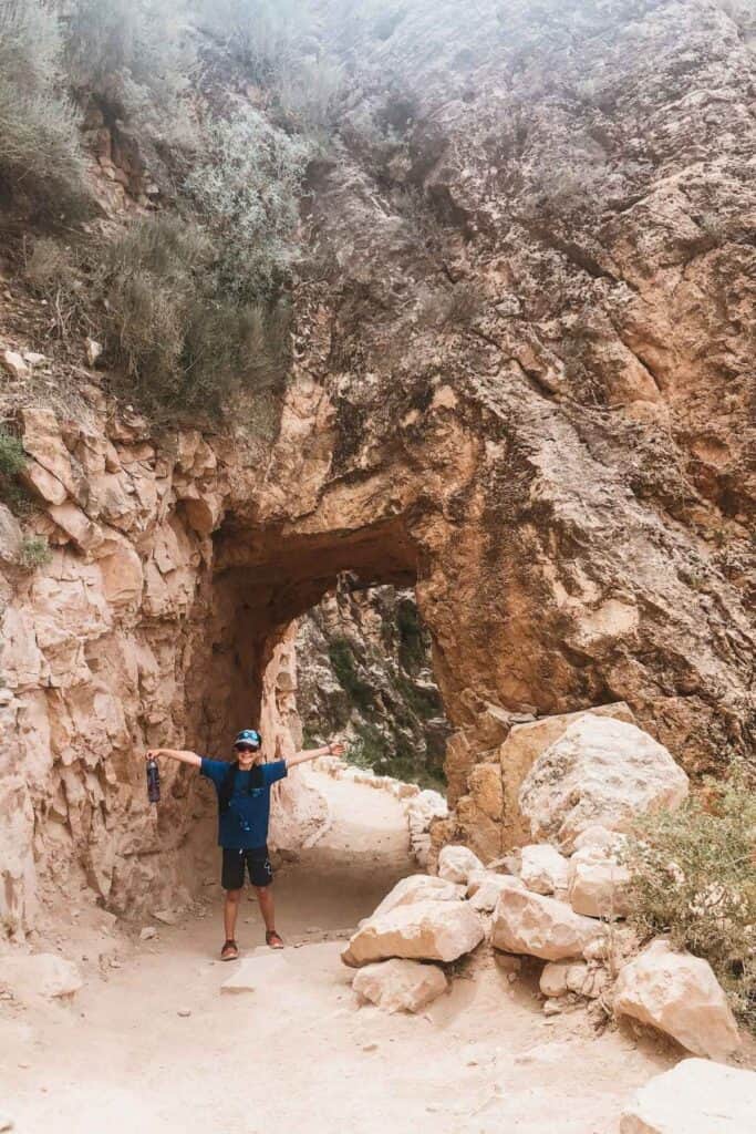 boy in blue shirt with arms stretched wide standing in front of a rock tunnel pass