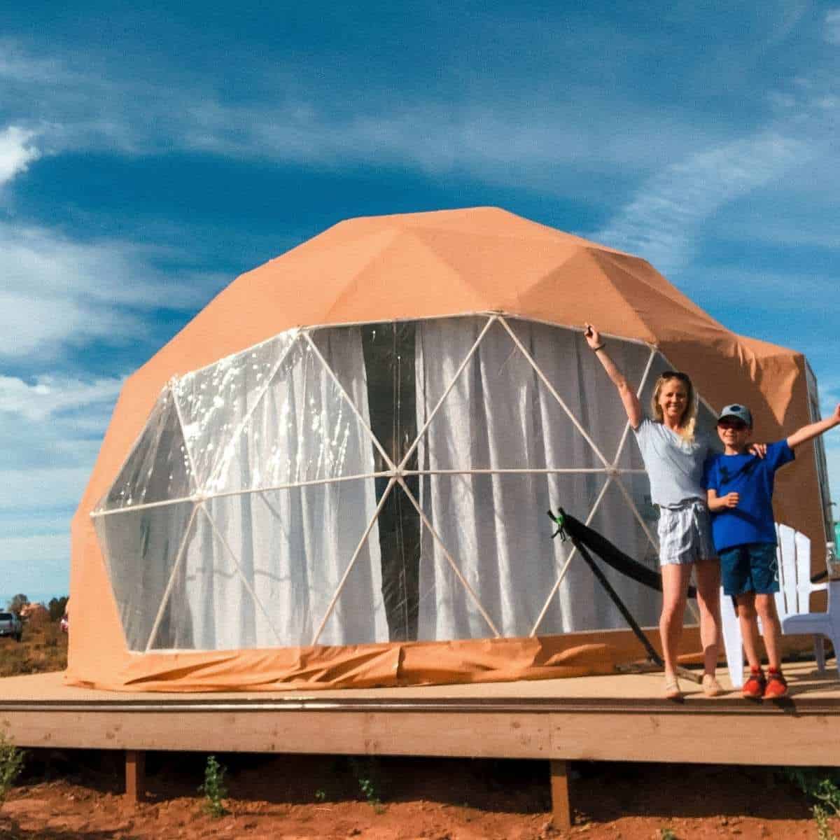 mother and son standing in front of a tan tent dome with blue sky in background