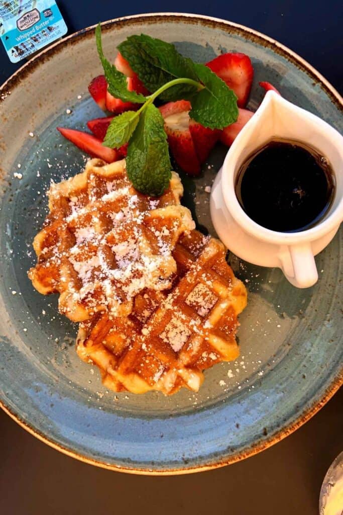 looking down on a blue tinted plate with 2 small belgian waffles, strawberries and a white pouring glass of maple syrup
