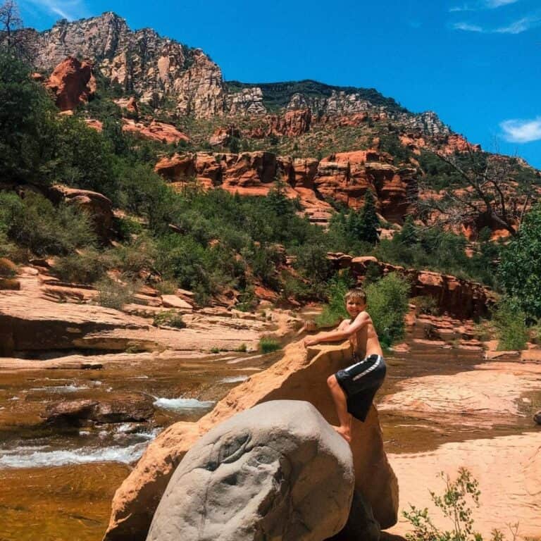 Boy in black shorts climbing on a red rock with river and red cliff rocks in background