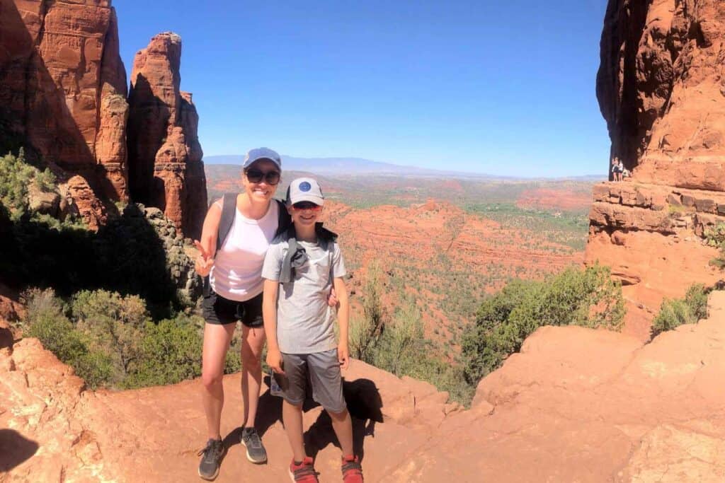 mother and son wearing hats and sunglasses standing on top of a red rock mountain with a valley below in the background 