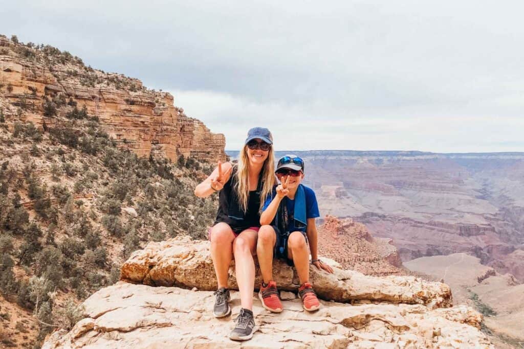 mom and son wearing hats sitting on a rock giving the peace sign with their fingers with a mountain valley behind them