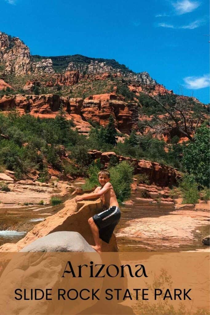 boy on top of red rock in black shorts in the middle of a river with red cliff background and blue sky