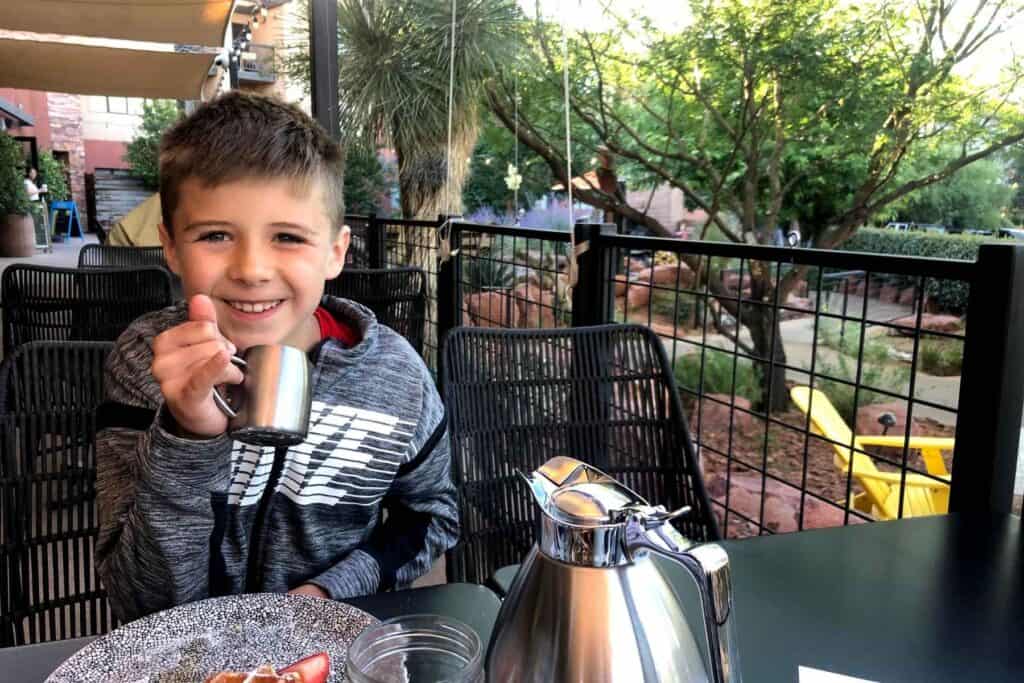 boy sitting at a breakfast table holding up his syrup cup for waffles with his pinky up