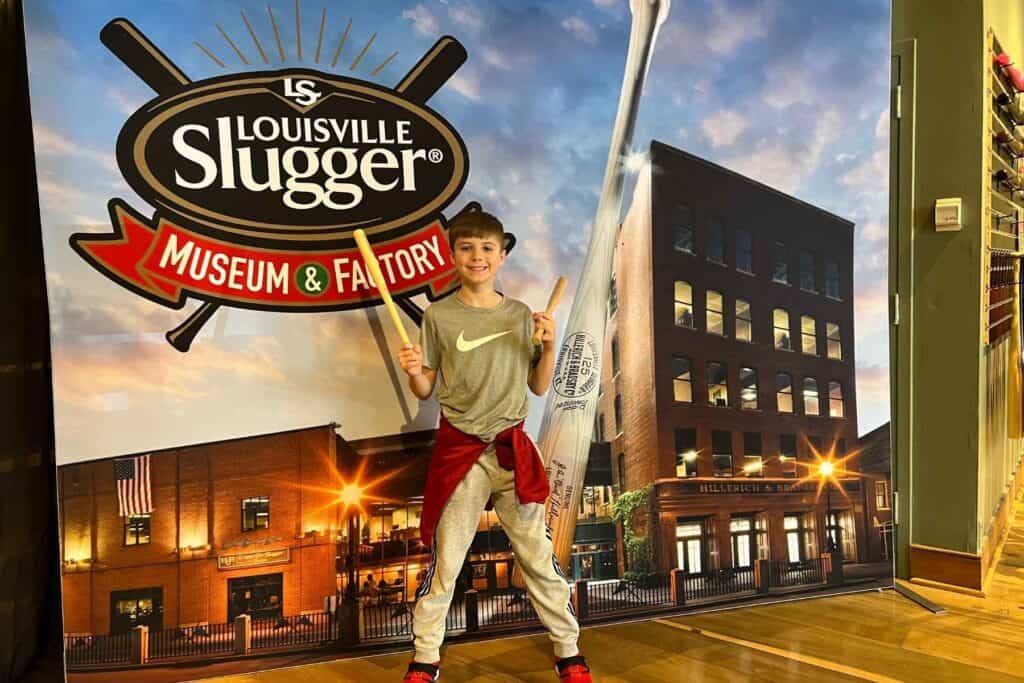 boy holding two miniature baseball bats in from of Louisvill Slugger Museum poster 