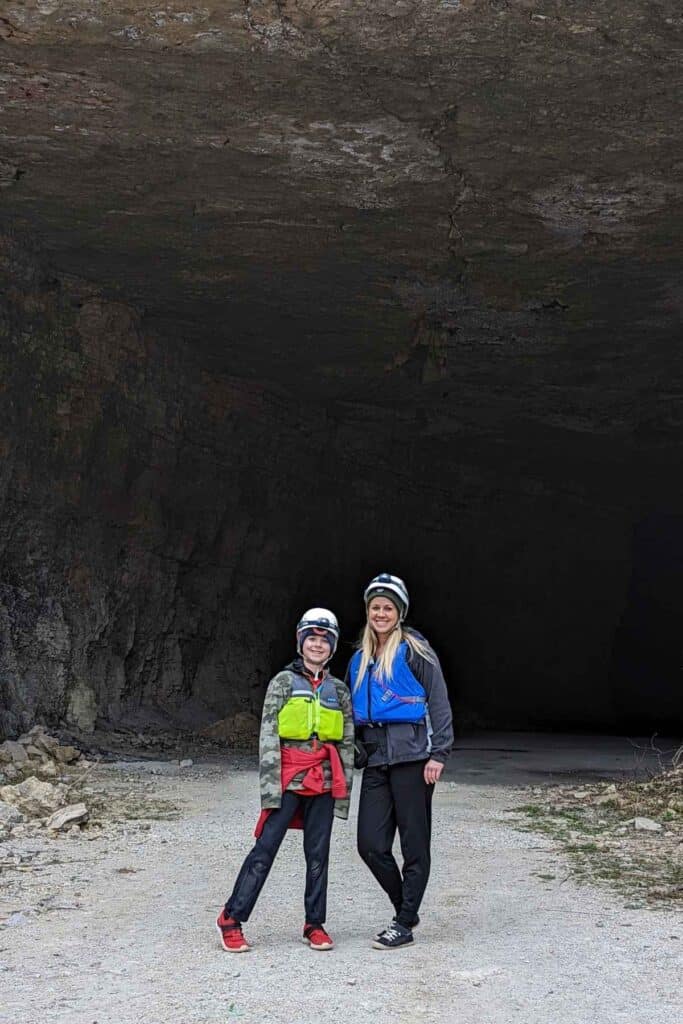 Mom and son standing in front of a cavern hole wearing helmets and life jackets