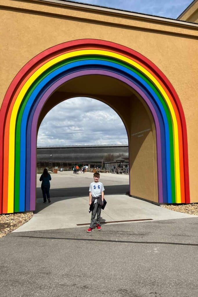 boy in gray short sleeves shirt with gray pants standing under a rainbow entrance to the ark exhibit 
