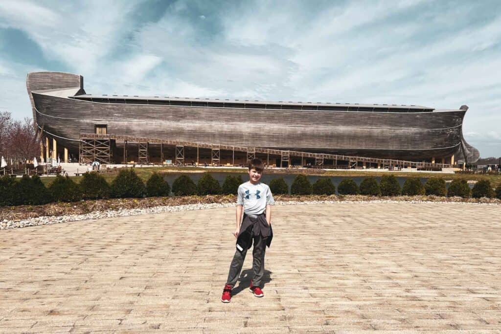 a boy in a grey shirt standing on a tan brick road in front of a big wooden boat