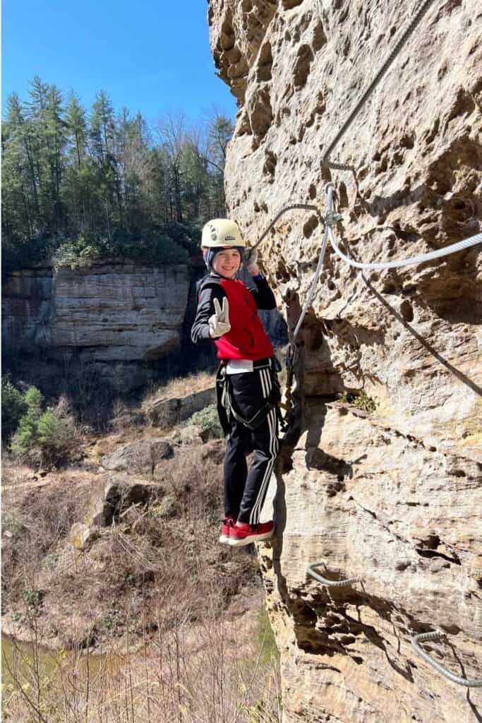 Boy in white and black outfit  climbing on side of a cliff with a white helmet giving a peace sign with hand