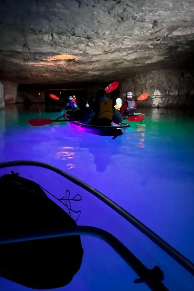 front of a crystal clear kayak looking at other glowing kayak in a dark cave