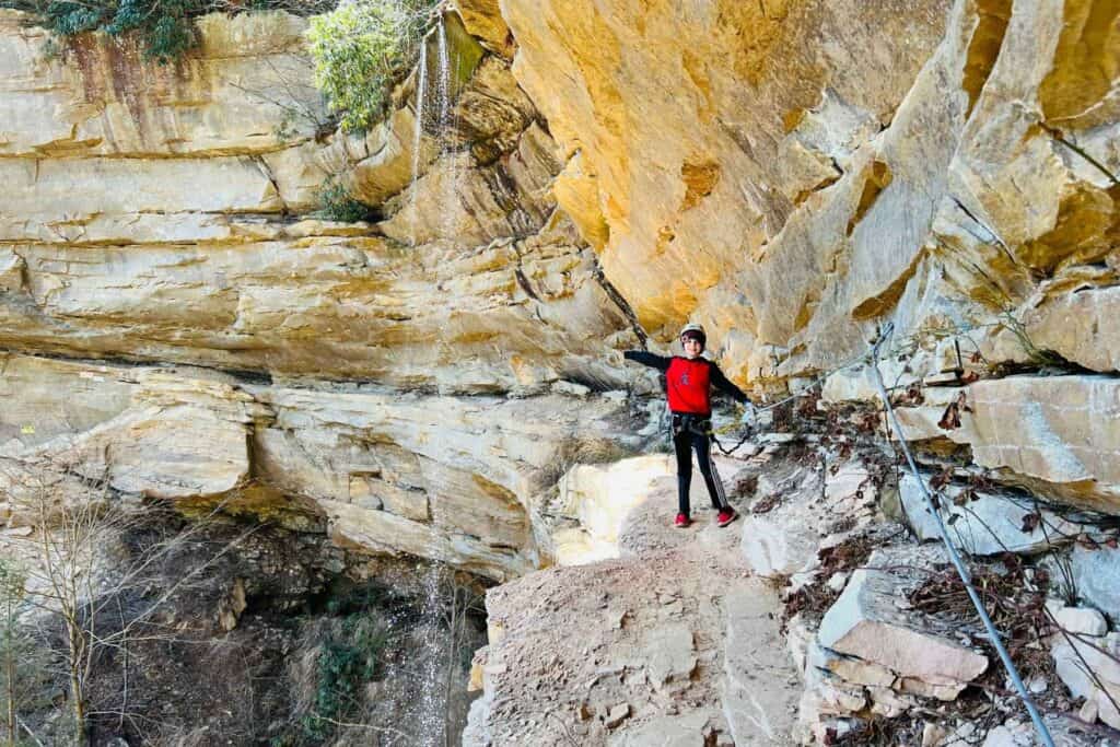 boy in a red and black outfit on the side of a cliff behind a waterfall