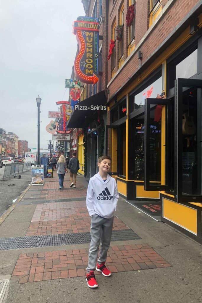 boy in white shirt and grey pants standing on broadway street with all the signs behind him