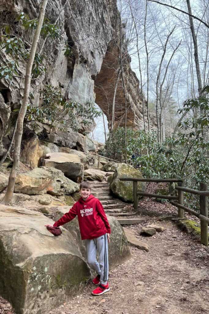 boy in red sweatshirt and grey sweatpants standing around rock formation with a natural rock bridge behind him. One of Kentucky's top outdoor attractions. 