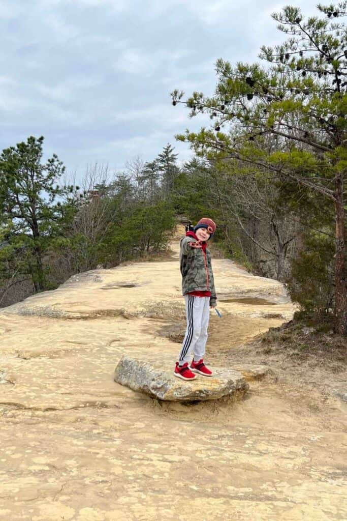 boy standing on top of a nartural sandstone arch in a camo coat giving the peace sign with his hand at natural bridge state park and hemlock lodge