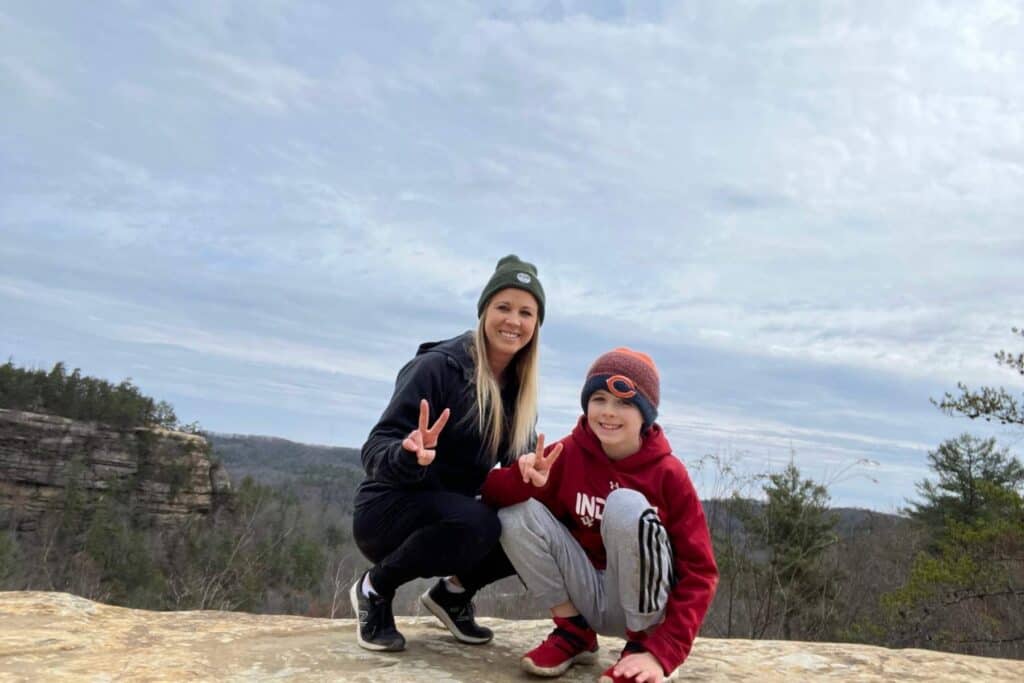 mom and son squatting giving a peace sign with hands  with a forest behind them
