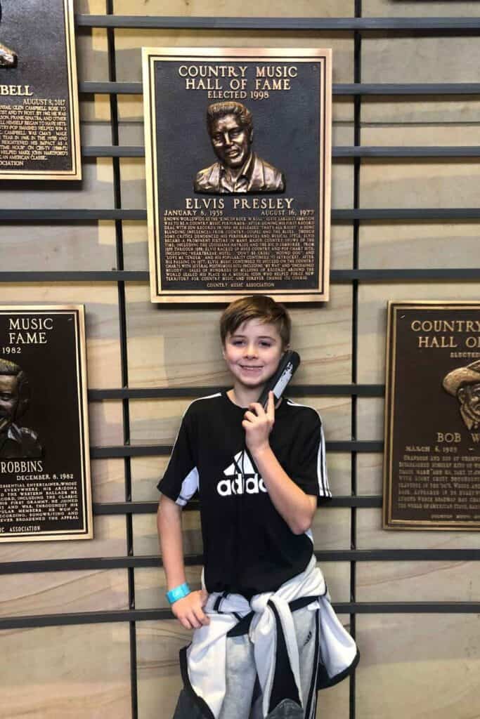 boy in black shirt standing in front of Elvis Presly's plaque at the Country Music Hall of Fame listening to a recording headset 