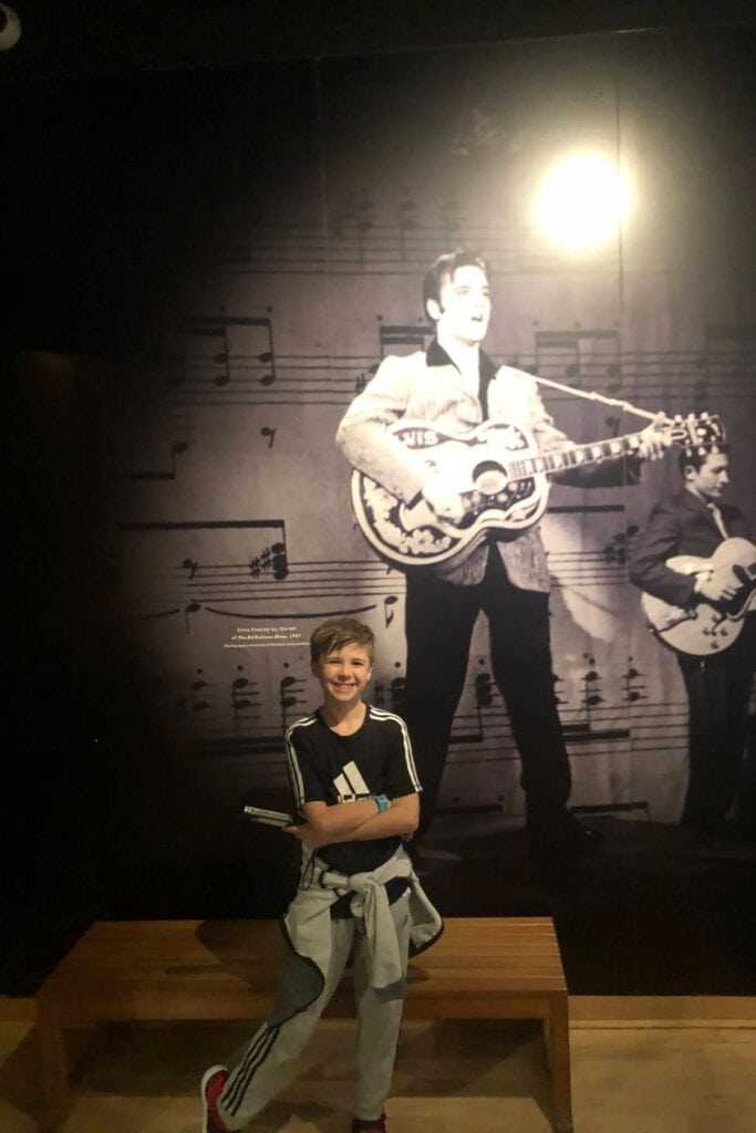boy standing in front of wall with a poster of young Elvis Presley singing with his guitar 
