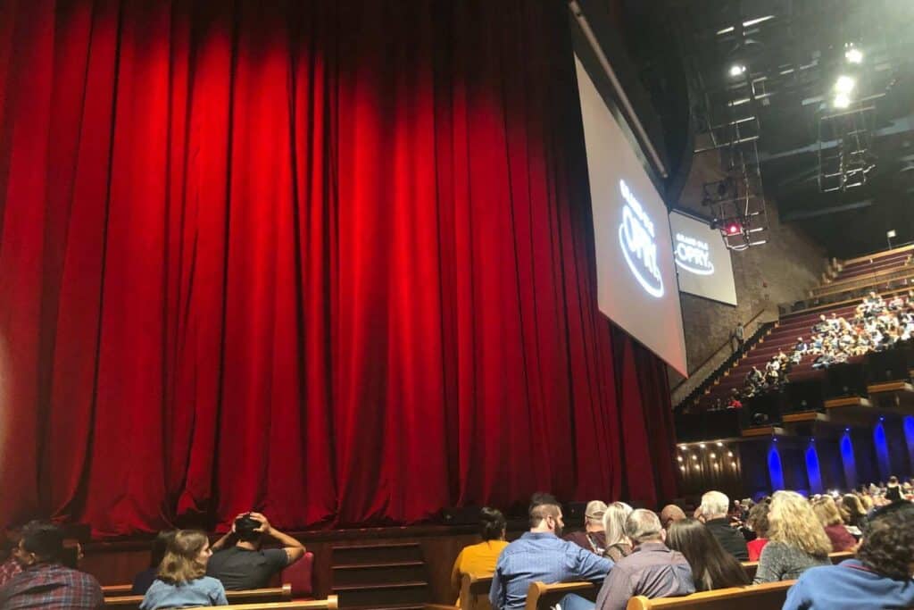 Red velvet curtains covering the stage of the Grand Ole Opry with people sitting in the wooden pews anticipating the upcoming show. 
