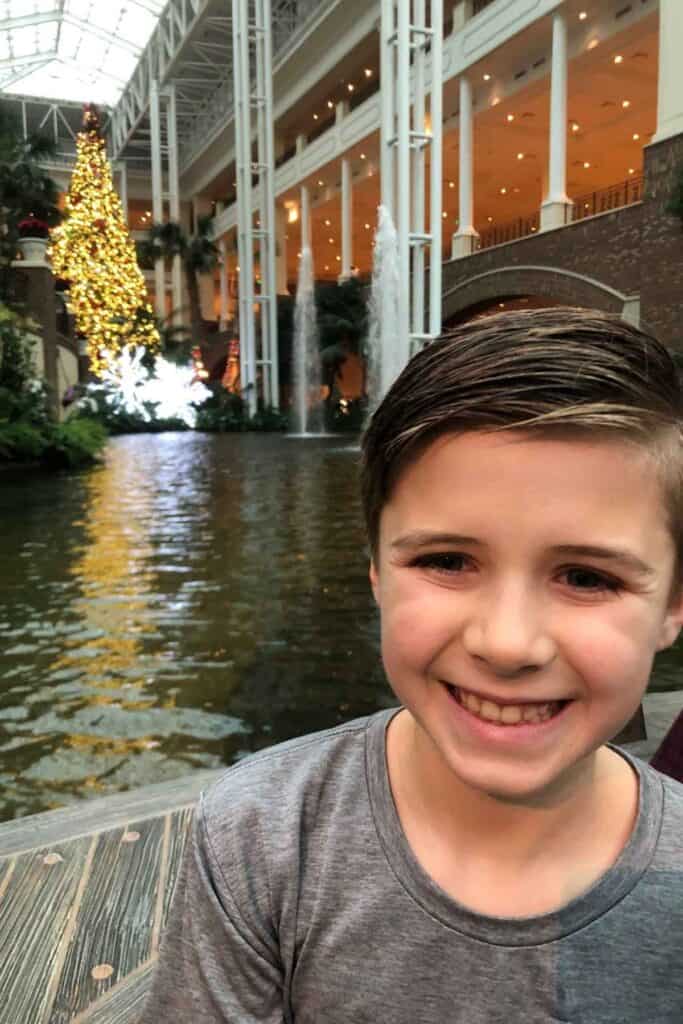 Young boy in a grey shirt enjoying a riverboat with Christmas tree lit up behind him at Gaylord Opryland Resort