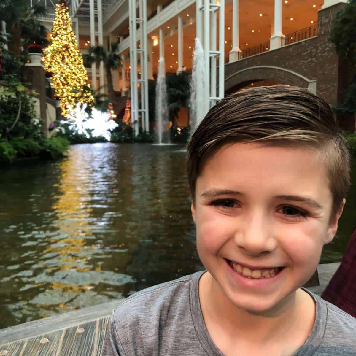 boy in grey shirt smiking with a river and a lit up christmas tree behind him