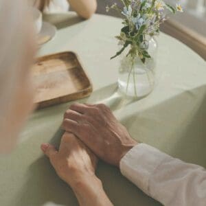 Elderly couple holding hands across a table with a small vase of flowers, symbolizing love, caregiving, and human connection