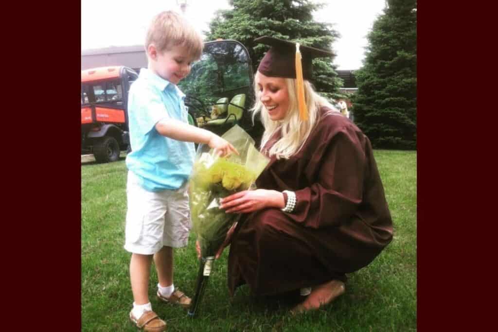 Mother in graduation cap kneeling beside her young son as he gives her flowers, capturing a heartfelt moment of love and connection